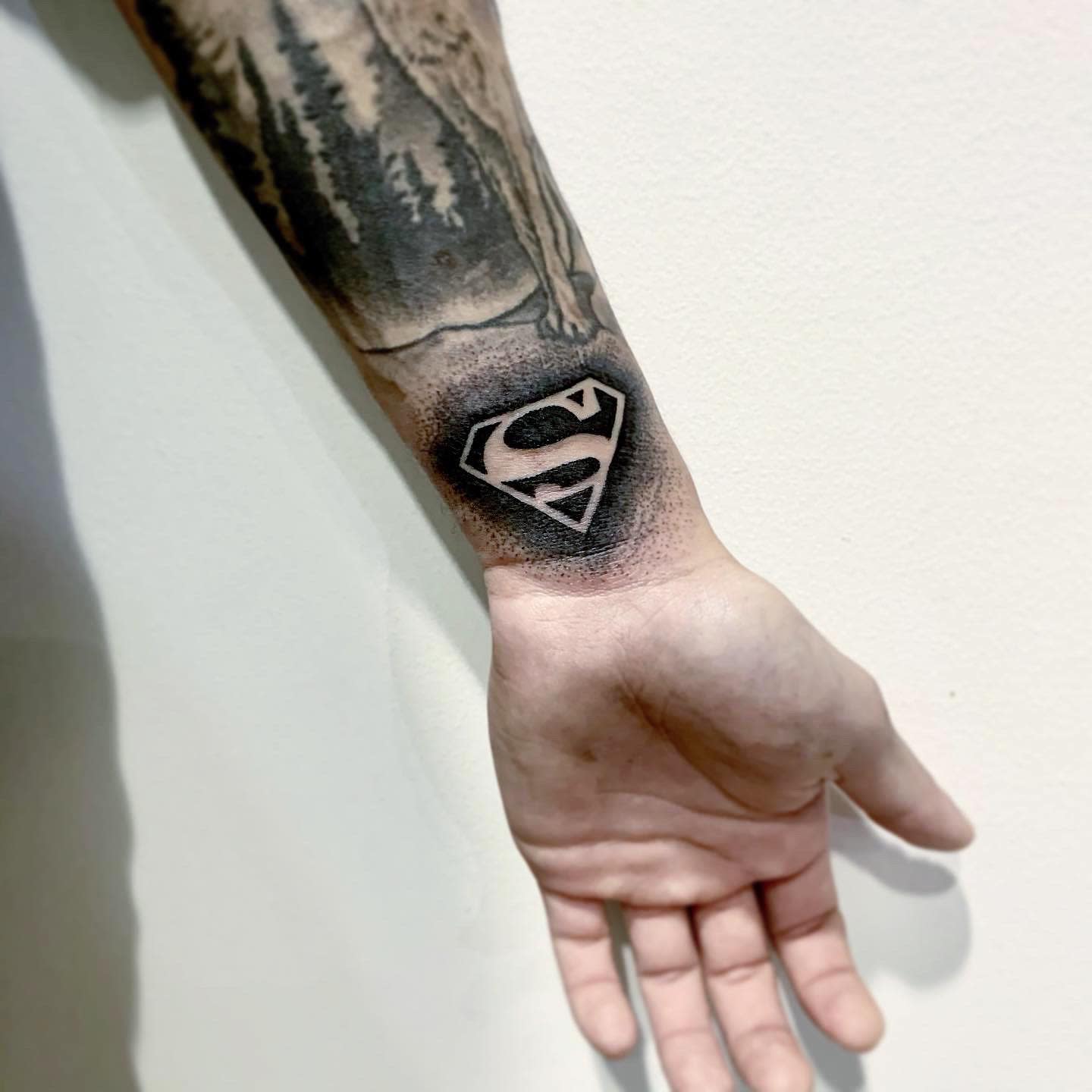 Wrist tattoo of Superman logo in black ink beneath a forest and wolf tattoo