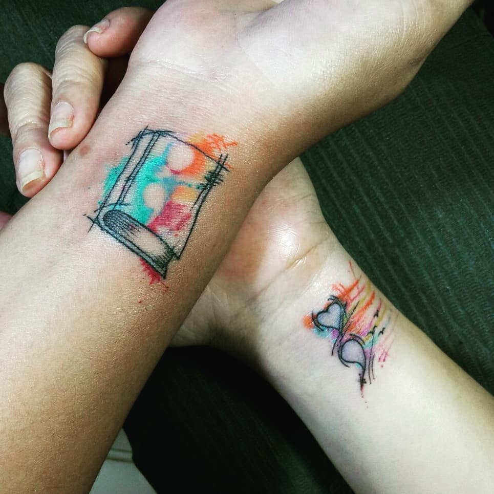 Two wrists with colorful watercolor-style tattoos; one of a book and the other of hearts