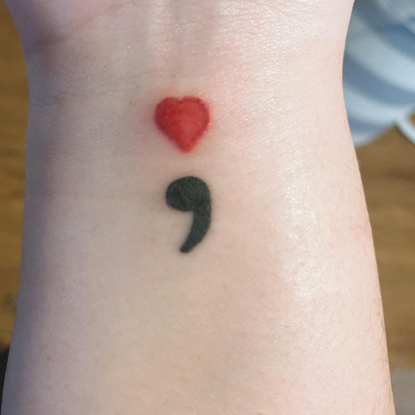 Tattoo of a red heart above a black semicolon on a wrist