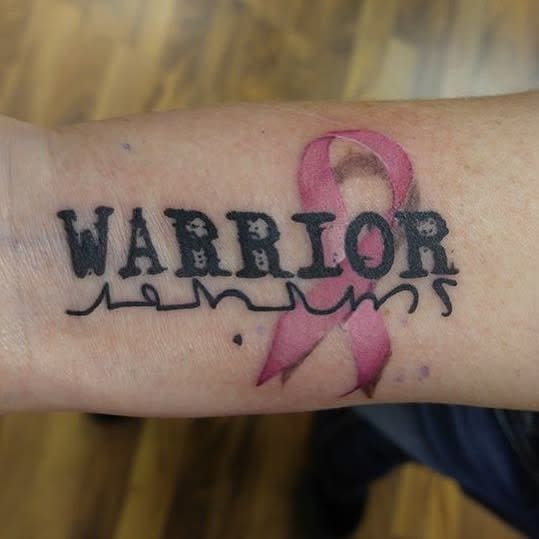 Tattoo of the word "WARRIOR" with a pink ribbon on an arm symbolizing breast cancer awareness
