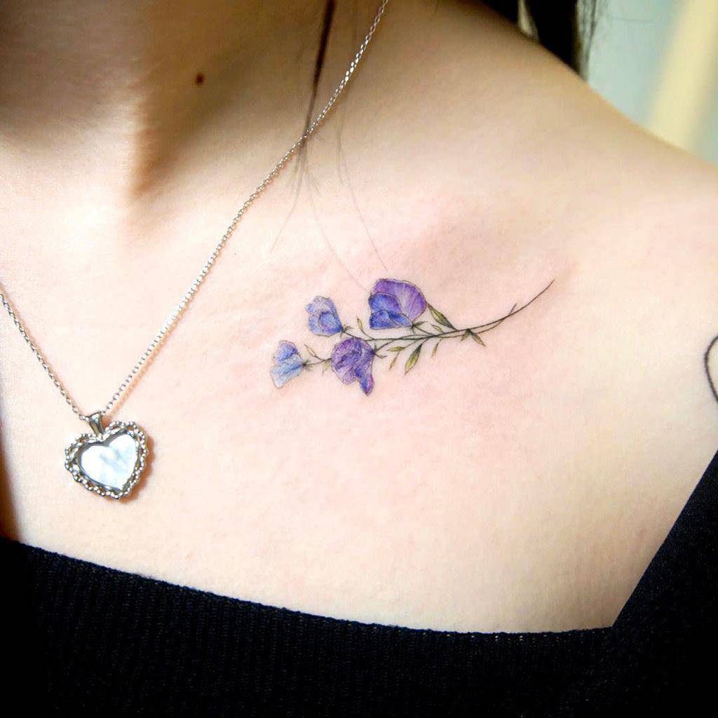 A minimal and elegant sweet pea flower tattoo in light shades of purple, adding a subtle touch to the shoulder