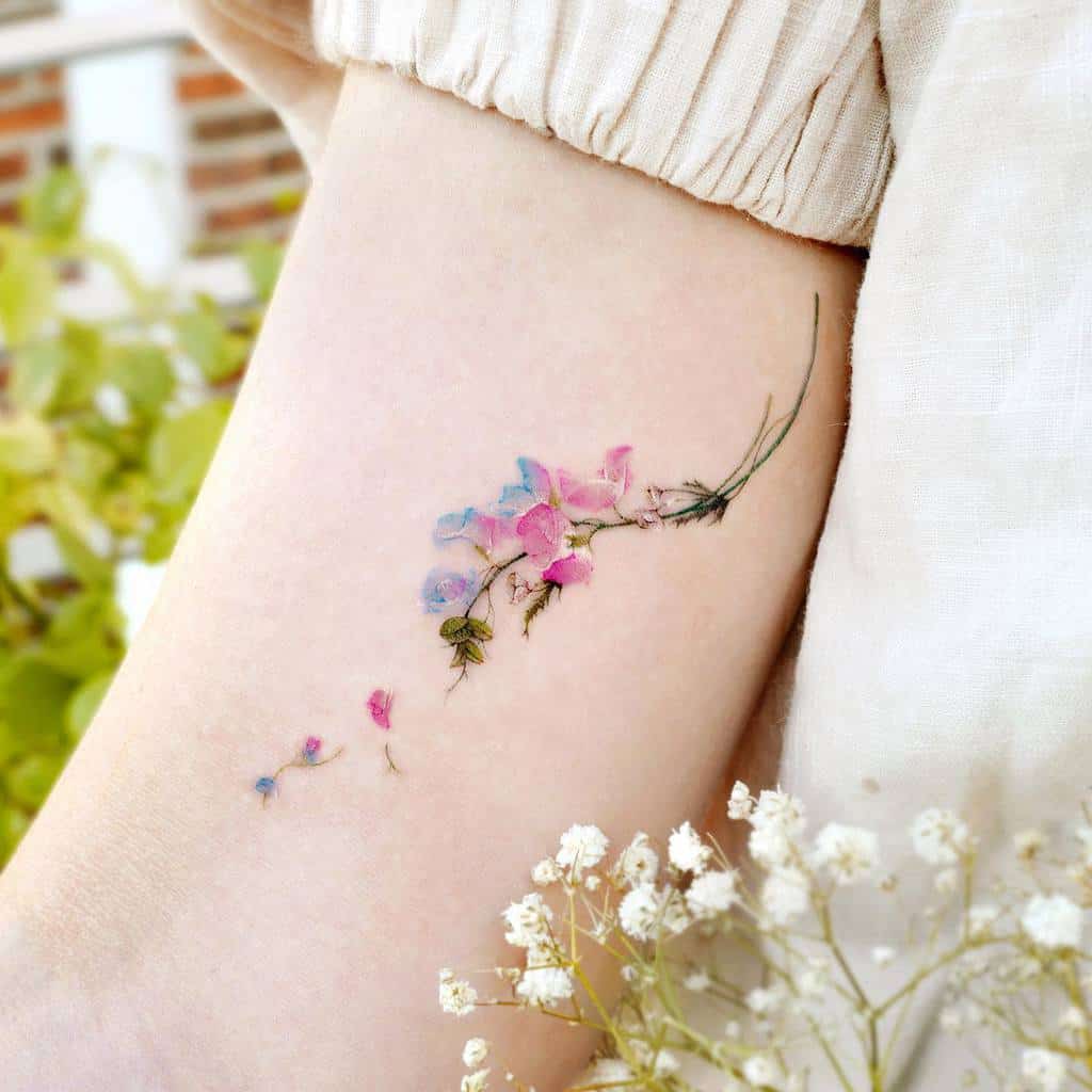 Delicate pastel sweet pea flower tattoo on the upper arm, featuring soft pink and blue hues