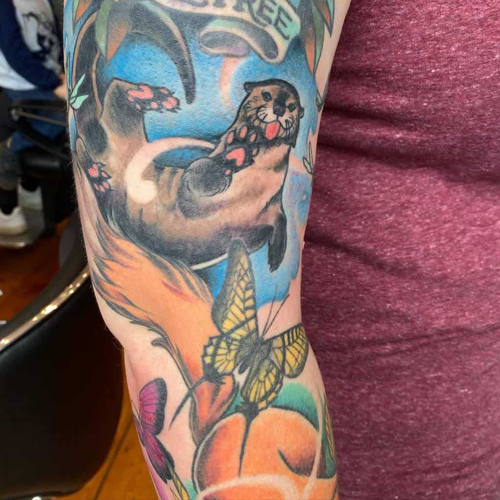 Vibrant tattoo of a cheerful otter with colorful surroundings, including butterflies, flowers, and a blue water-inspired background