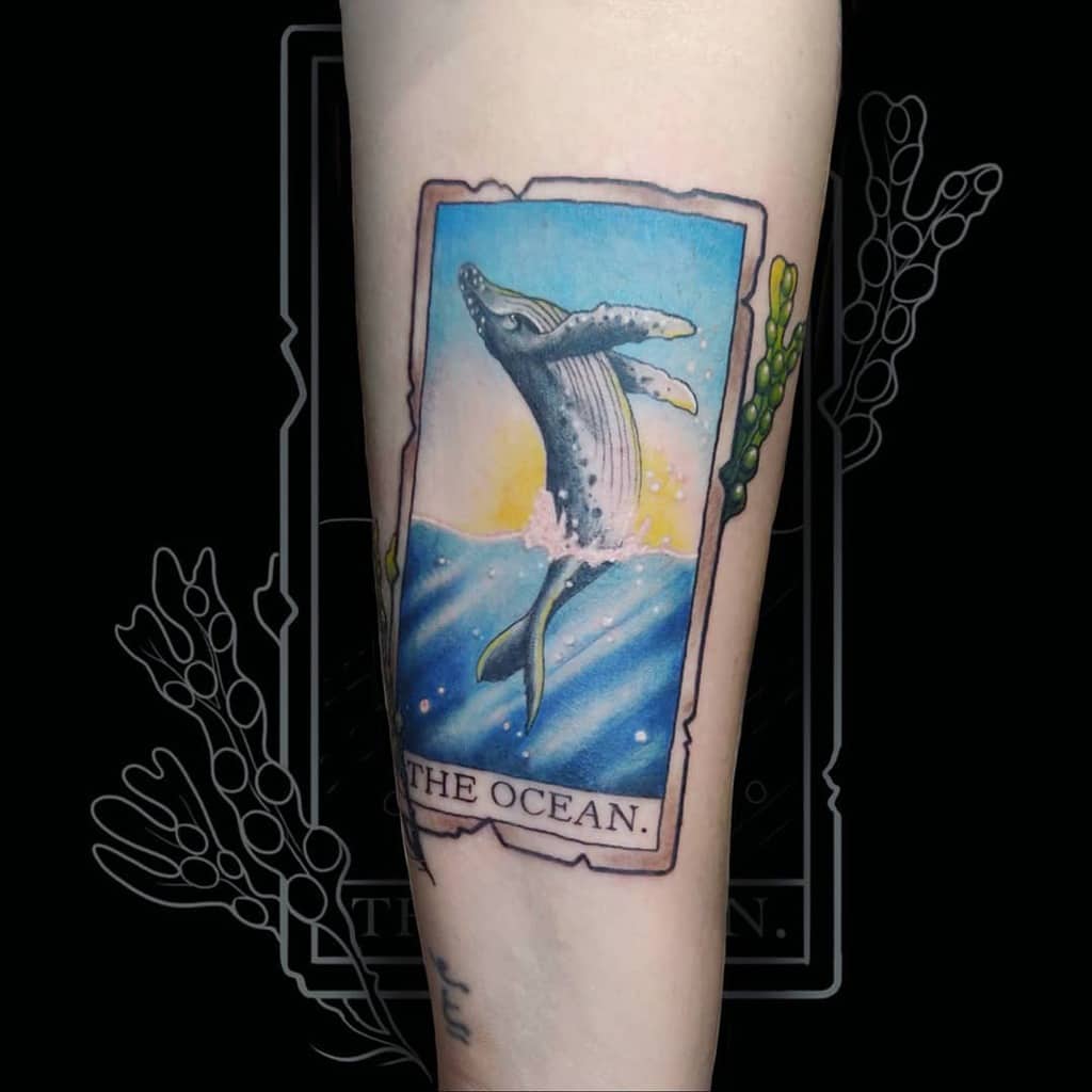Colorful tarot-style tattoo featuring a breaching whale against a sunset, framed within a card design with 'The Ocean' inscribed at the bottom and beans on the side