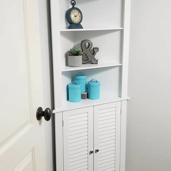Tall white corner bathroom cabinet with open shelves and lower shutter-style doors.