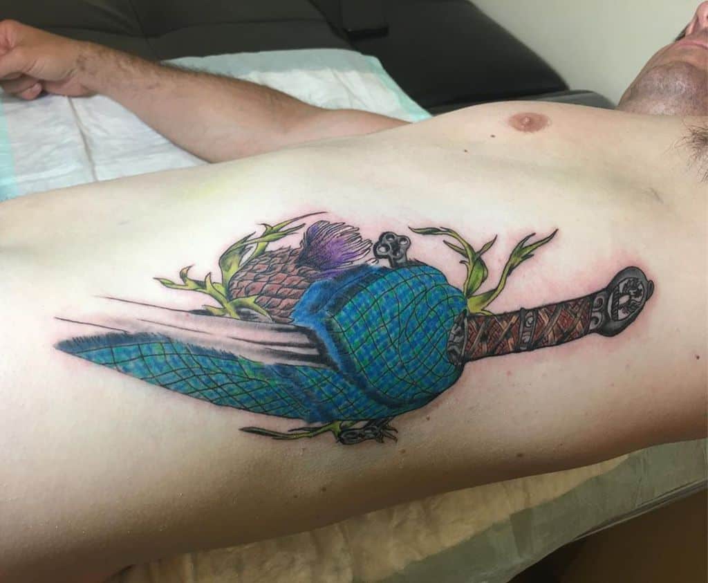 Vibrant tattoo featuring a Scottish dirk with a tartan pattern sheath, adorned with thistle and leaves
