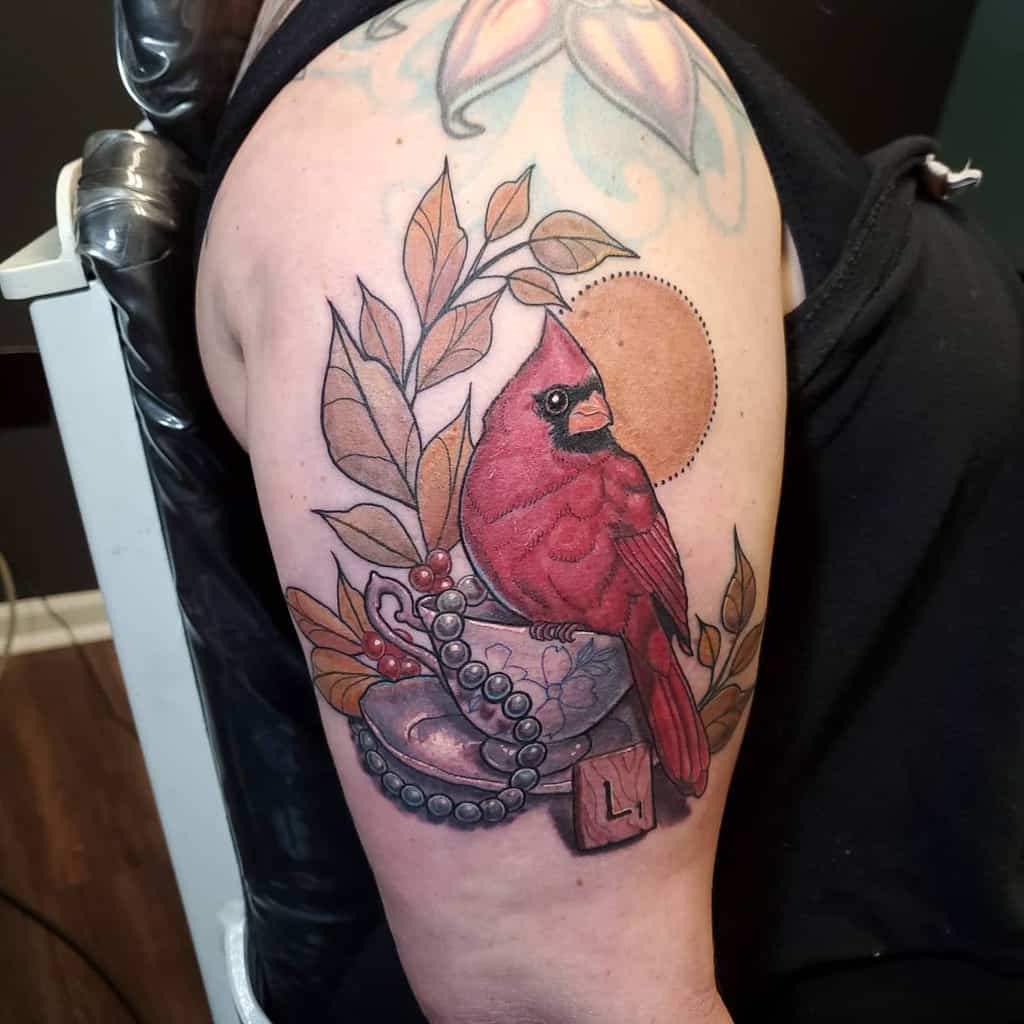 Colorful tattoo of a red cardinal perched on a vintage floral teacup resting on a saucer, surrounded by leaves, a pearl necklace, a wooden tag with "L1," and a sun