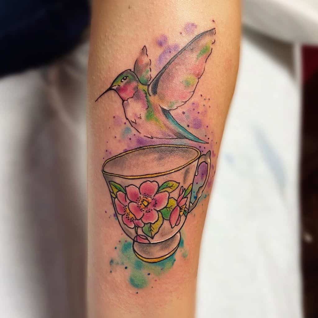 Colorful watercolor-style tattoo of a hummingbird hovering above a floral teacup, surrounded by vibrant splashes of blue, purple, and green