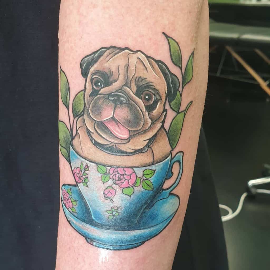 Colorful tattoo of a pug in a blue teacup with pink flowers and green leaves, resting on the matching saucer, surrounded by green leaves
