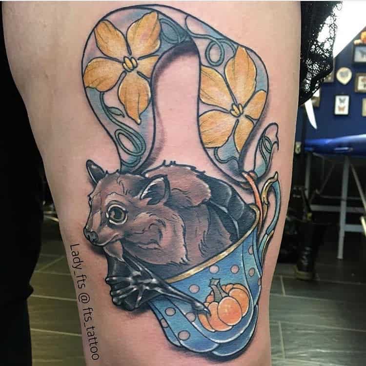 Colorful tattoo of a bat hanging from a blue teacup with polka dots and a pumpkin design, wrapped in a ribbon with yellow flowers