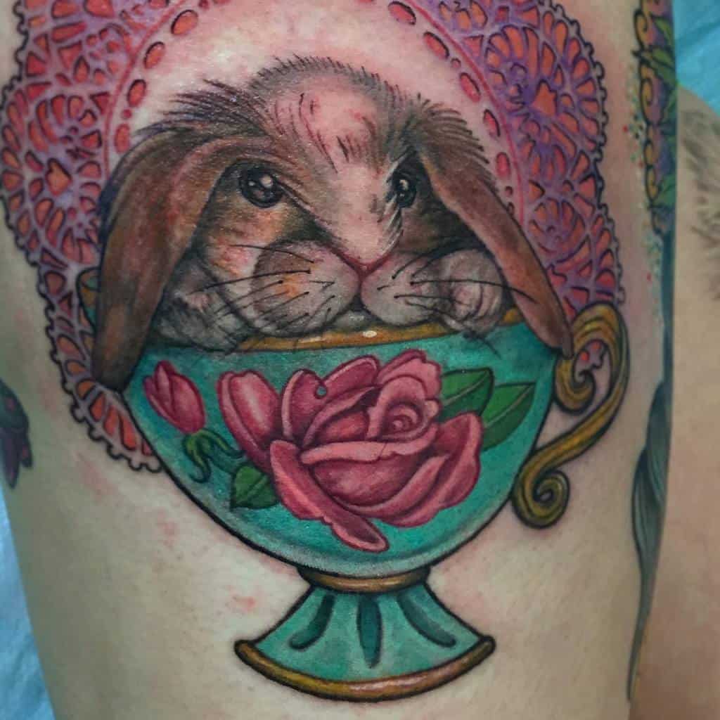 Colorful tattoo of a lop-eared bunny resting inside an ornate teacup with a floral rose design, set against a decorative lace background