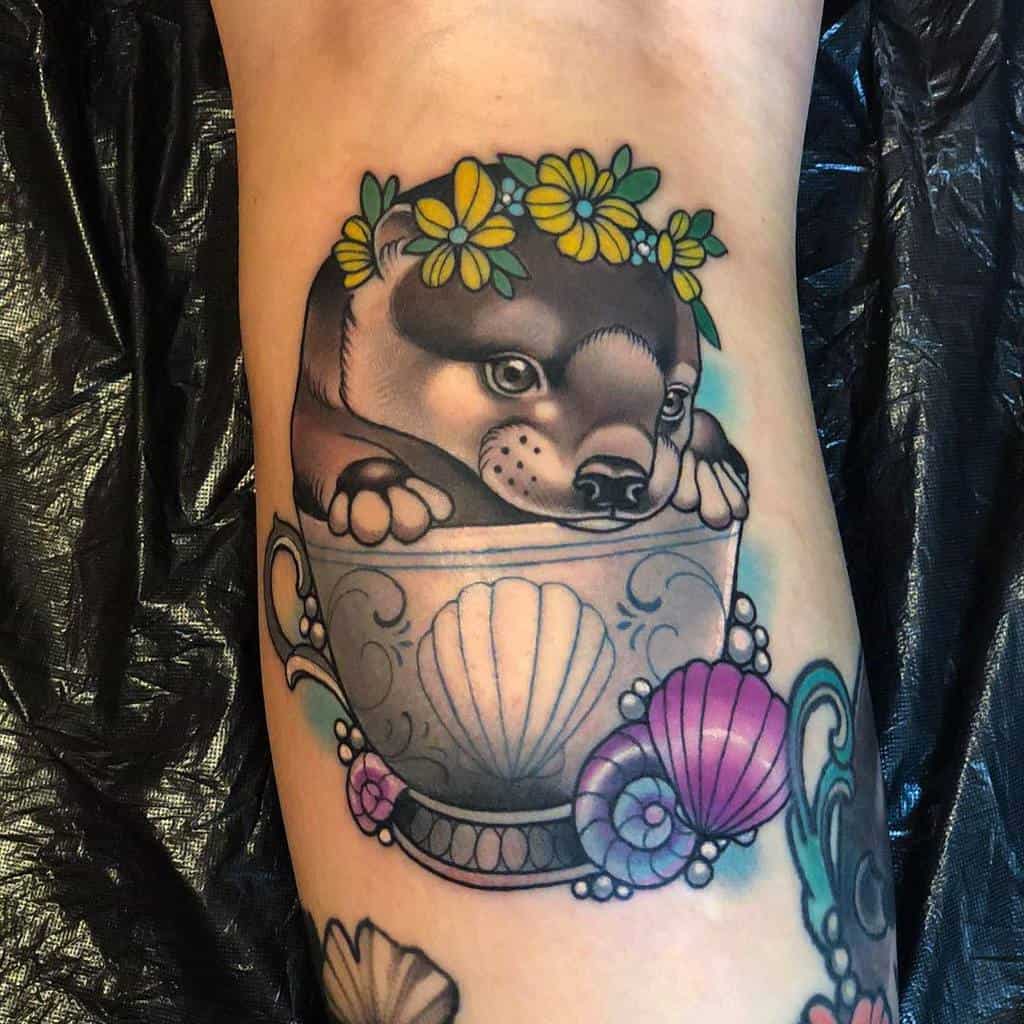 Colorful tattoo of a puppy with a yellow flower crown resting inside an ornate teacup with seashell and swirl designs, surrounded by pink and blue shells