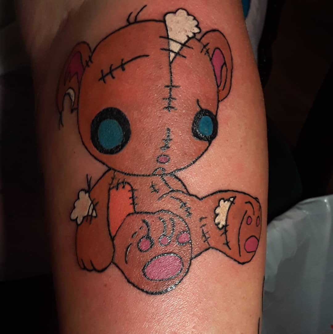 Tattoo of a cute, patched-up teddy bear with blue eyes and pink paws on skin