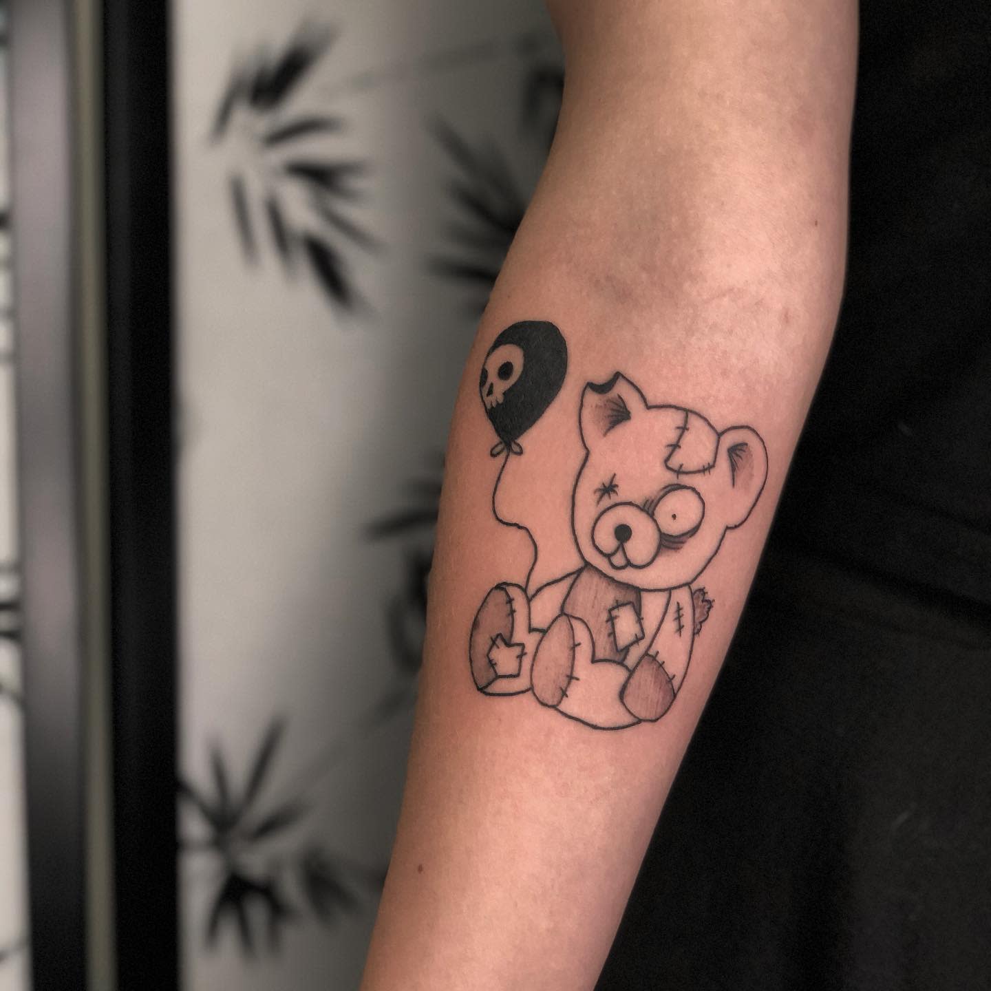 Tattoo of a stitched teddy bear holding a skull balloon on a forearm