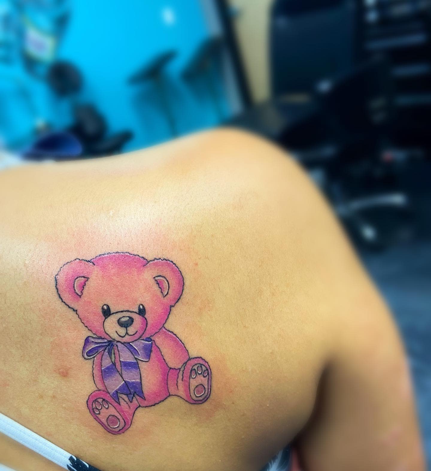 A pink teddy bear tattoo with a blue ribbon on the left shoulder