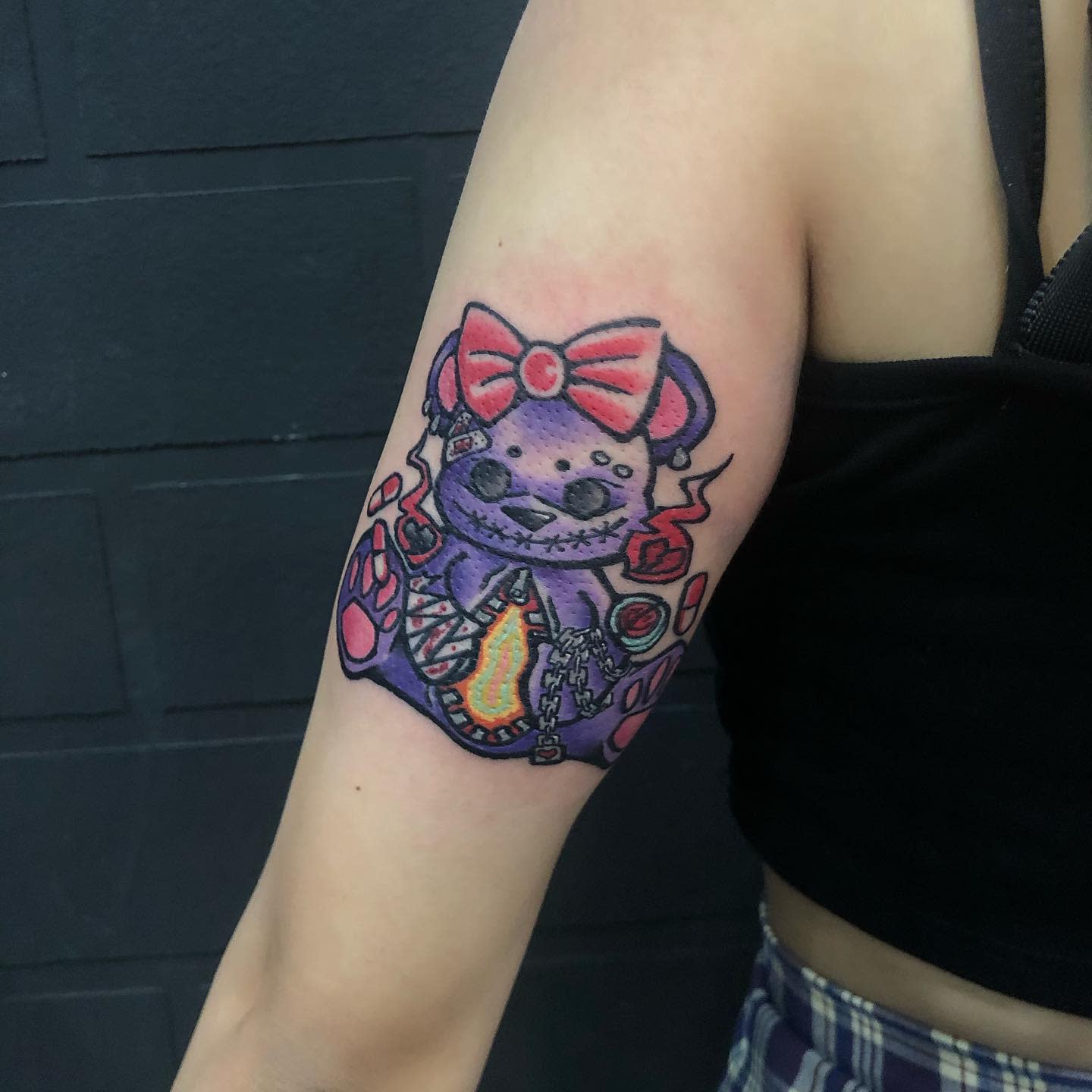 A colorful tattoo of an evil teddy bear with a red bow on its head, on an upper arm