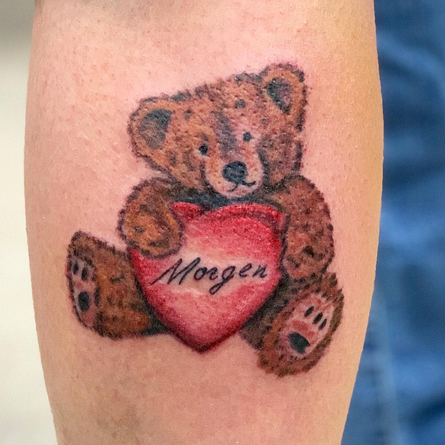 A tattoo of a teddy bear holding a red heart with "Morgen" written on it, inked on an arm