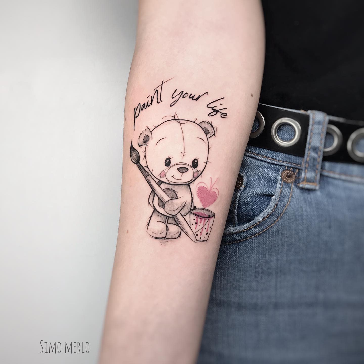 Tattoo of a cute teddy bear holding a paintbrush with "paint your life" text on an arm