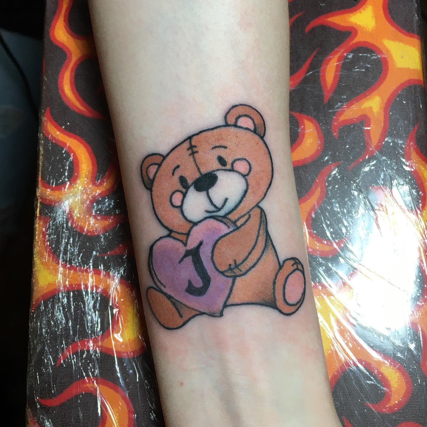 Tattoo of a cute teddy bear holding a heart with a "J" on the arm against a fiery background