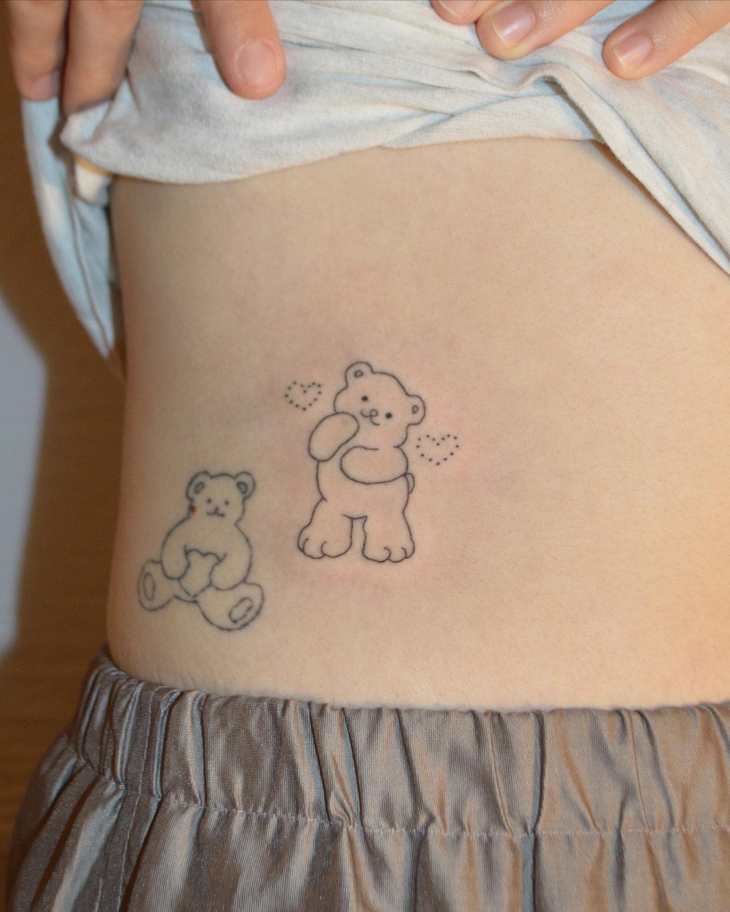 Tattoo of two outlined teddy bears on the side of a torso, near the waistline