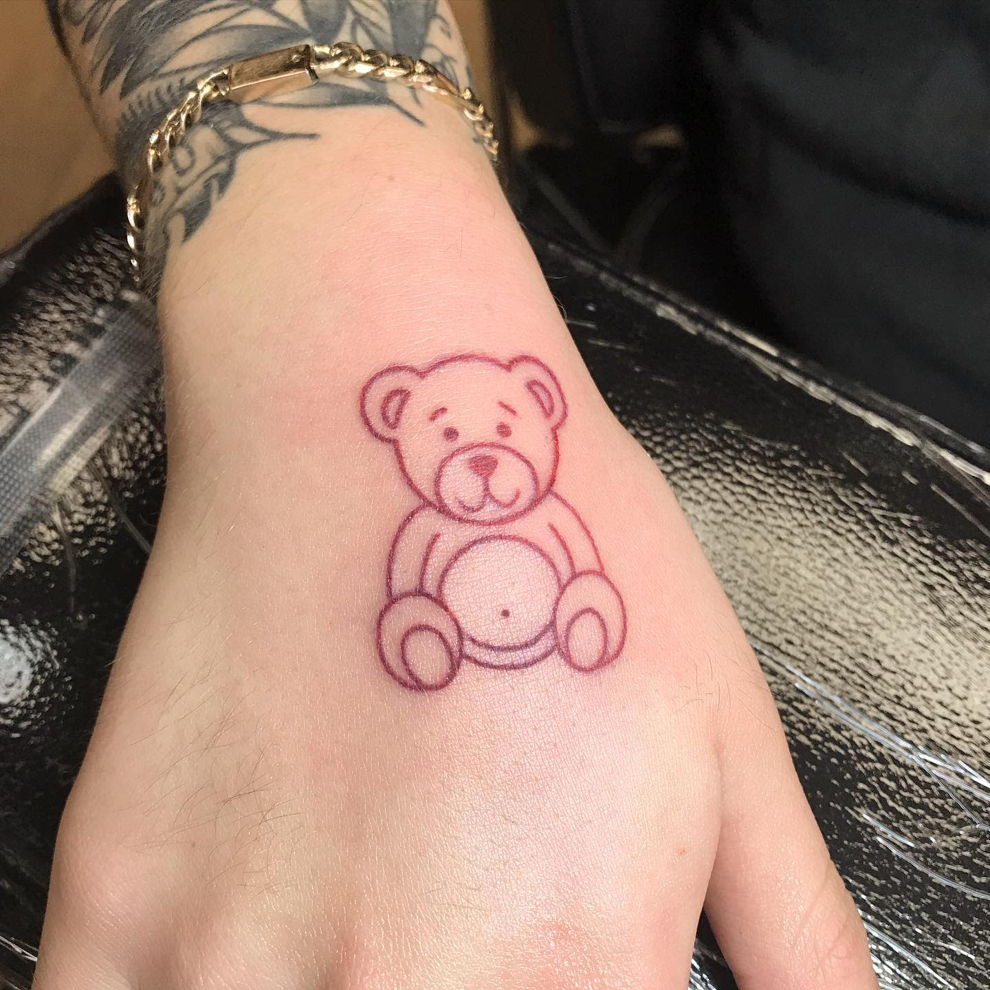 Tattoo of a simple, outlined teddy bear on the top of a hand