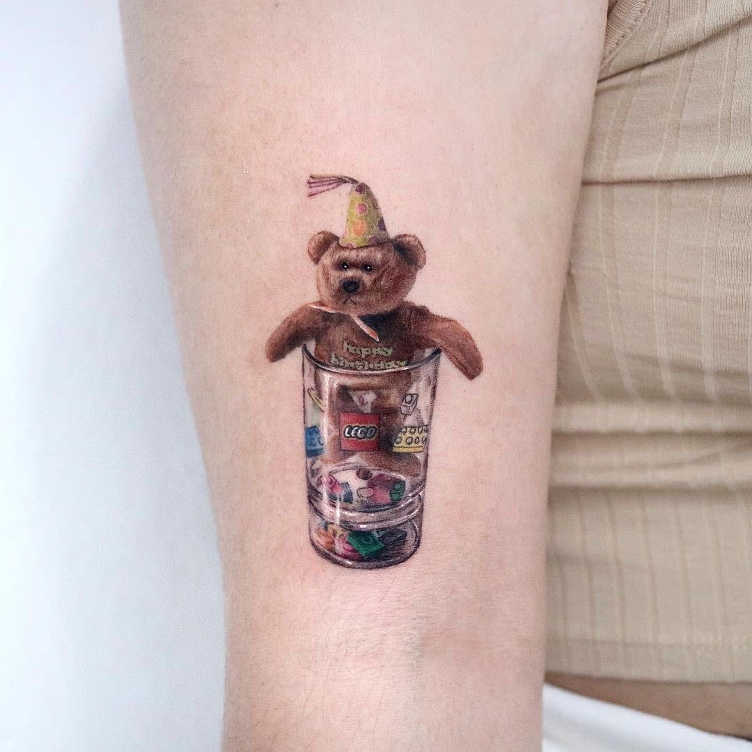 Realistic Teddy Bear Tattoo -inkedbooking Tattoo of a teddy bear in a party hat sitting in a cup filled with small colorful objects