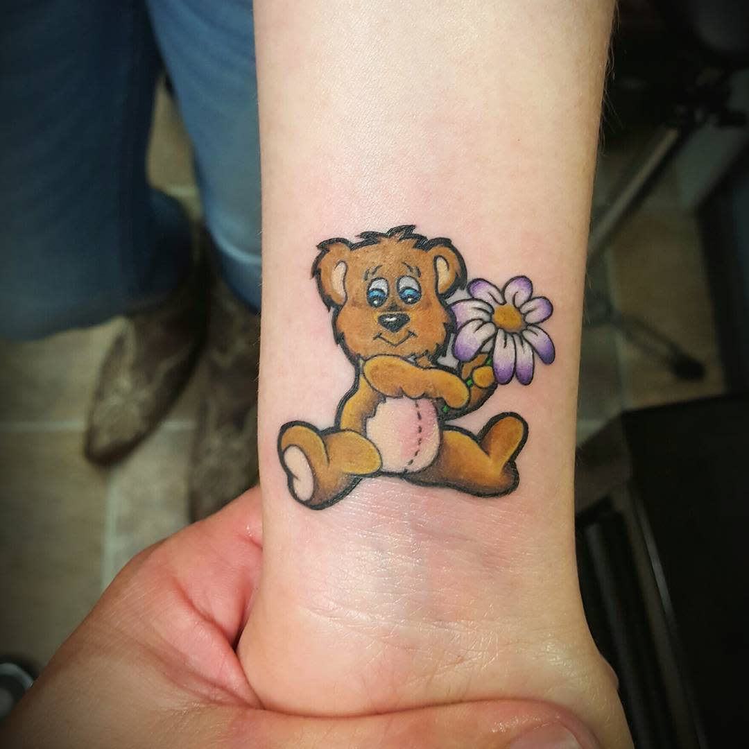 Tattoo of a cute teddy bear holding a purple flower on a forearm