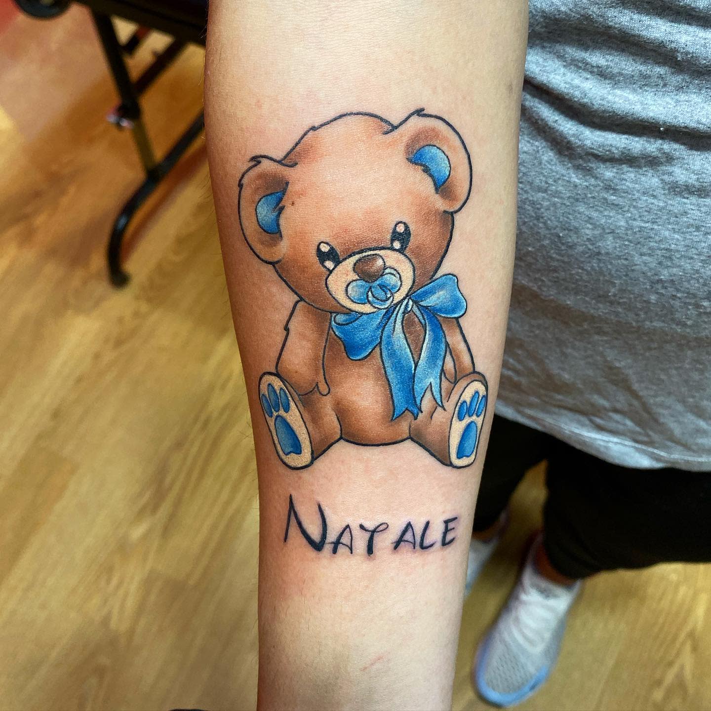 Tattoo of a blue-ribboned teddy bear with the name "Natalie" on a forearm