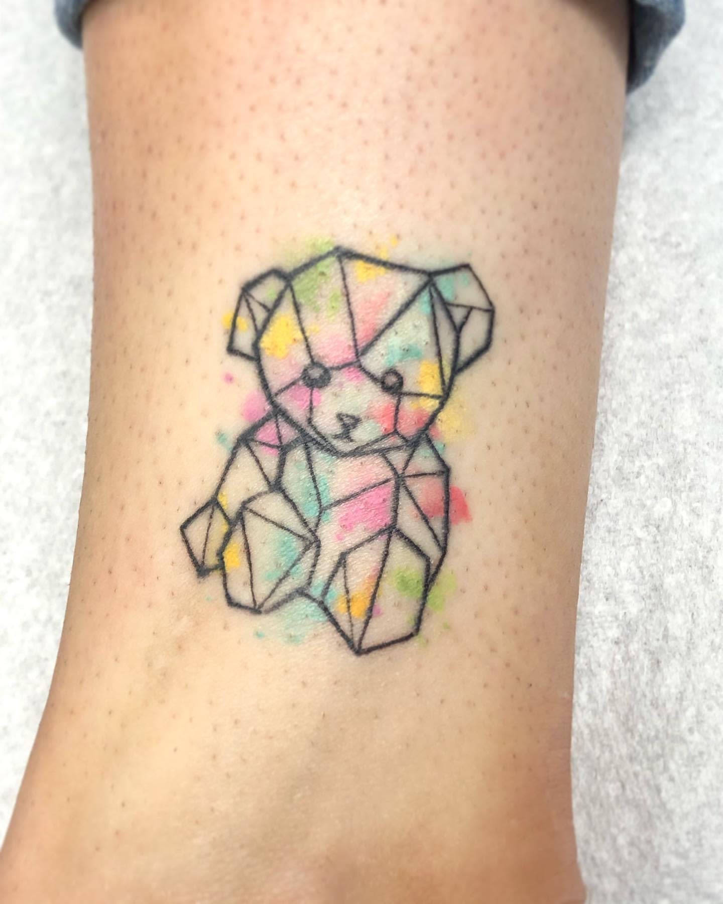 A geometric teddy bear tattoo with pastel watercolor accents on skin