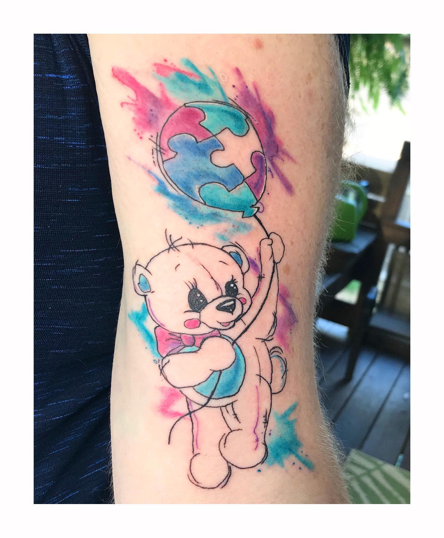 Tattoo of a teddy bear holding a colorful balloon with watercolor splashes on an upper arm