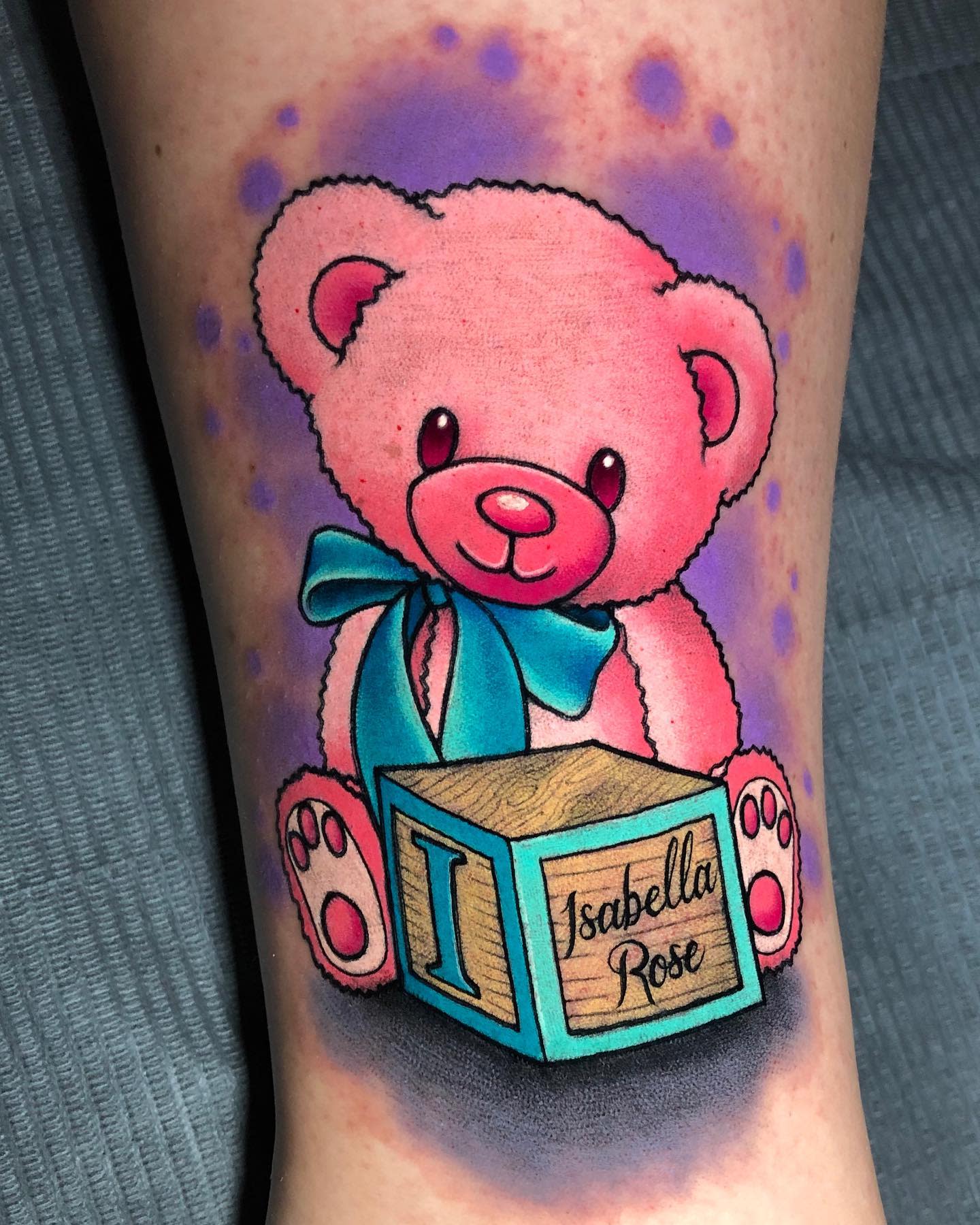 Tattoo of a pink teddy bear holding a blue ribbon and a block labeled "Isabella Rose"