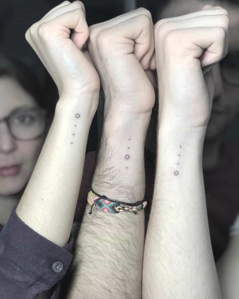 Three arms with matching dot and circle tattoos on their inner wrists, raised side by side