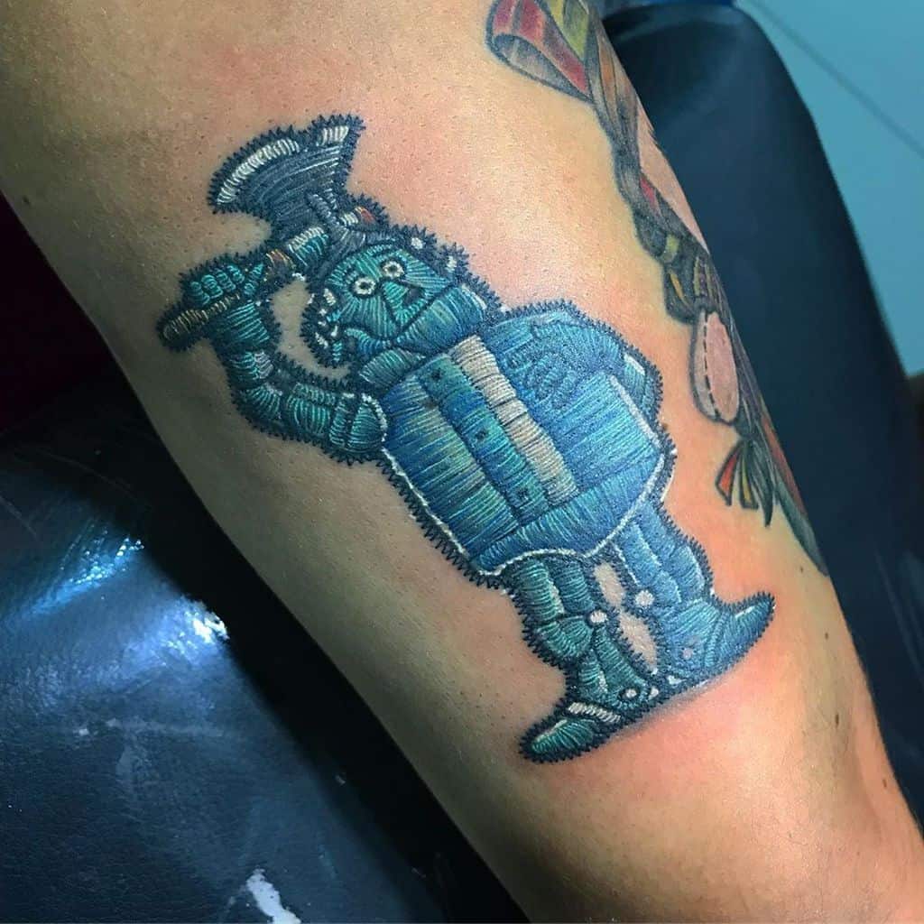 Realistic embroidered tattoo of a blue Tin Man with a thread-like texture, designed to resemble a sewn fabric patch.
