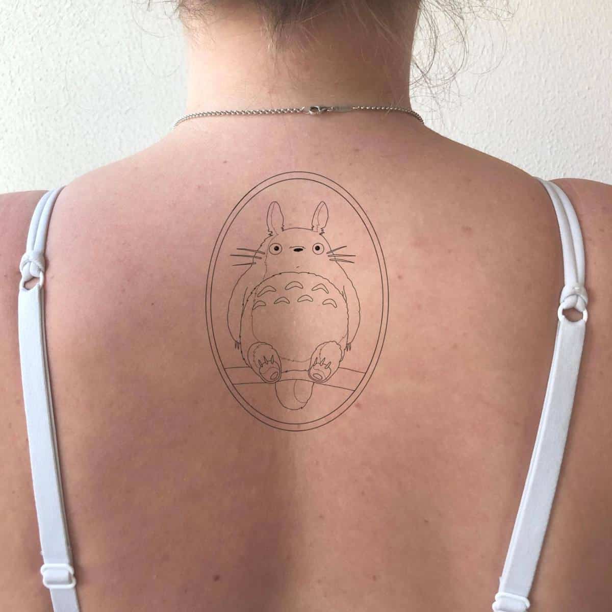 Simple line tattoo of Totoro from My Neighbor Totoro, centered on the upper back with an oval frame surrounding the character