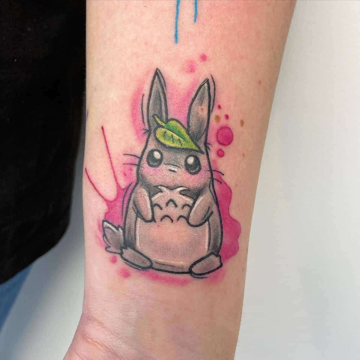 Colorful tattoo of Totoro from My Neighbor Totoro on the forearm, with a pink watercolor splash background and a green leaf on Totoro's head