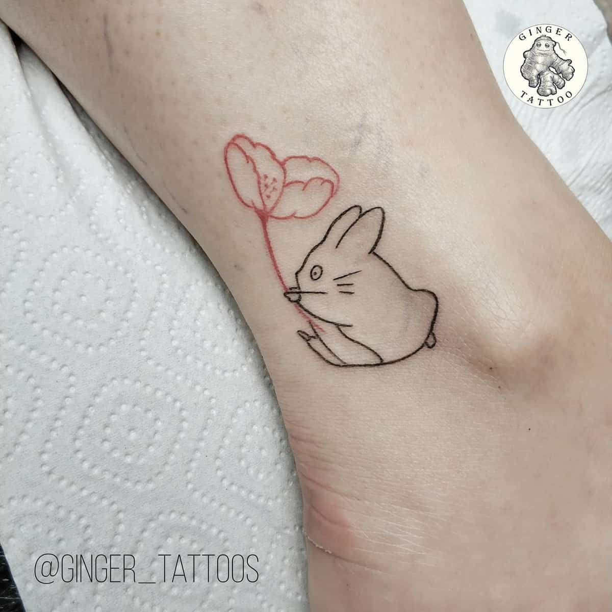 Minimalist ankle tattoo of a small Totoro holding a red flower. The design is simple, using fine lines to create a cute and delicate look