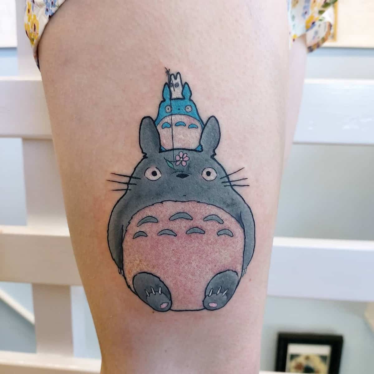 Thigh tattoo featuring Totoro with two smaller Totoro characters stacked on its head. The design is colorful and playful, with a pink flower on Totoro’s nose