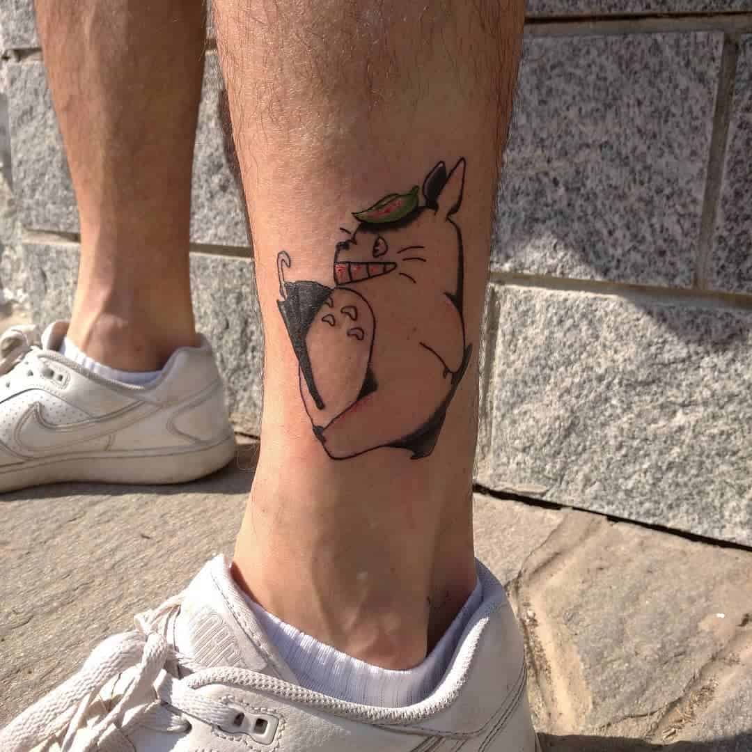 Ankle tattoo featuring Totoro holding an umbrella, with a leaf on its head. The design is minimalist, capturing Totoro’s playful expression and iconic features