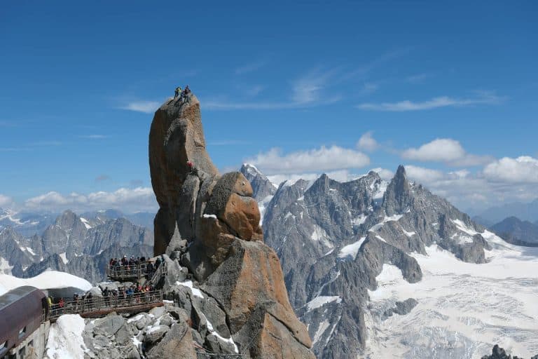 Tourists on a viewing platform and climbers on a towering granite spire, set against a vast panorama of snow-covered jagged mountains in the Alps