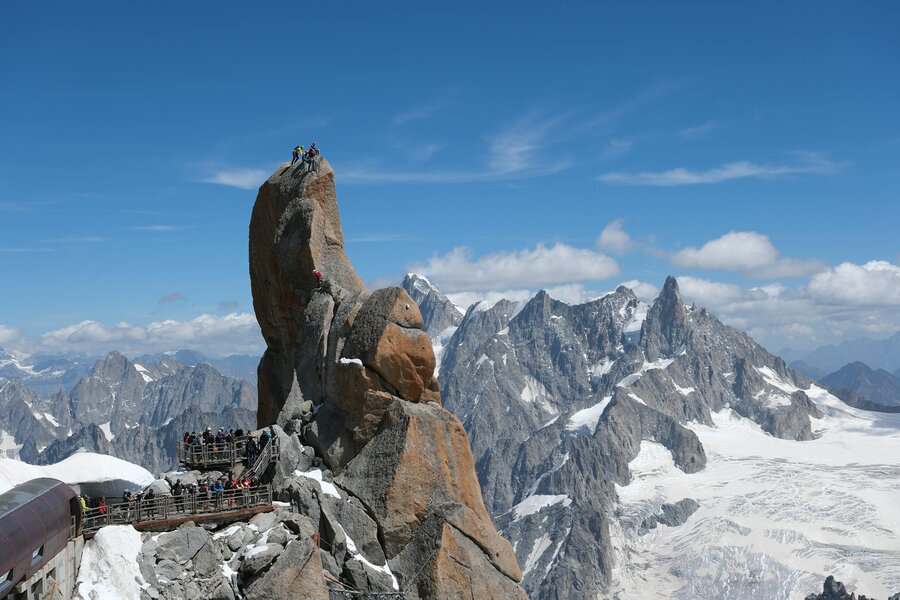 Tourists on a viewing platform and climbers on a towering granite spire, set against a vast panorama of snow-covered jagged mountains in the Alps