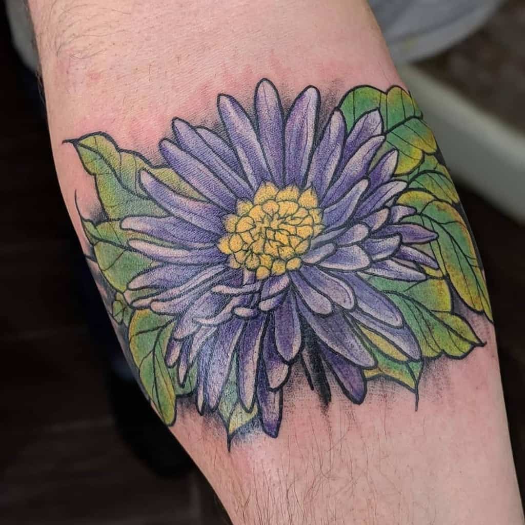 Tattoo of a purple aster with a yellow center and green leaves on a person's arm