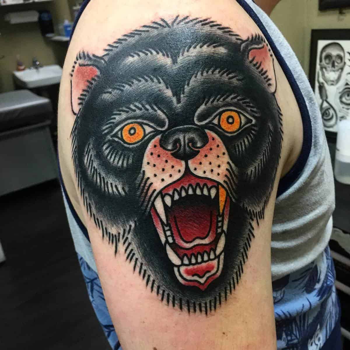 Tattoo of a roaring black bear with striking orange eyes inked on a person's upper arm