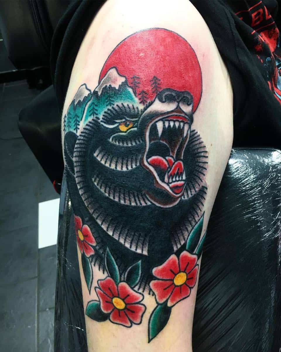 Tattoo of a roaring bear with a red sun, mountains, and vibrant red flowers on an arm