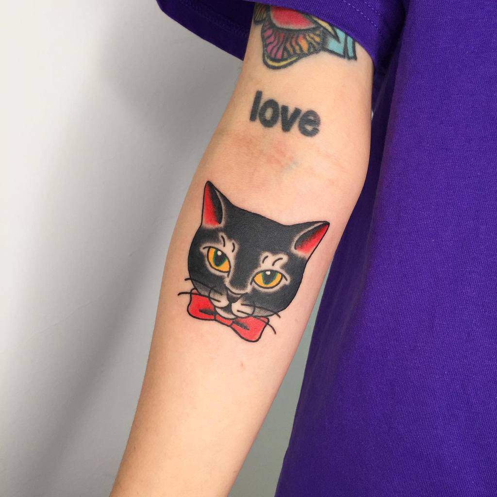 Tattoo of a sleek black cat with piercing yellow eyes and a bold red bow tie, artfully inked on a person's forearm