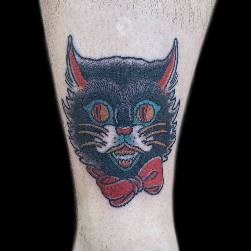 A tattoo of a fierce black cat with piercing orange eyes and a stylish red bow tie on a person's skin