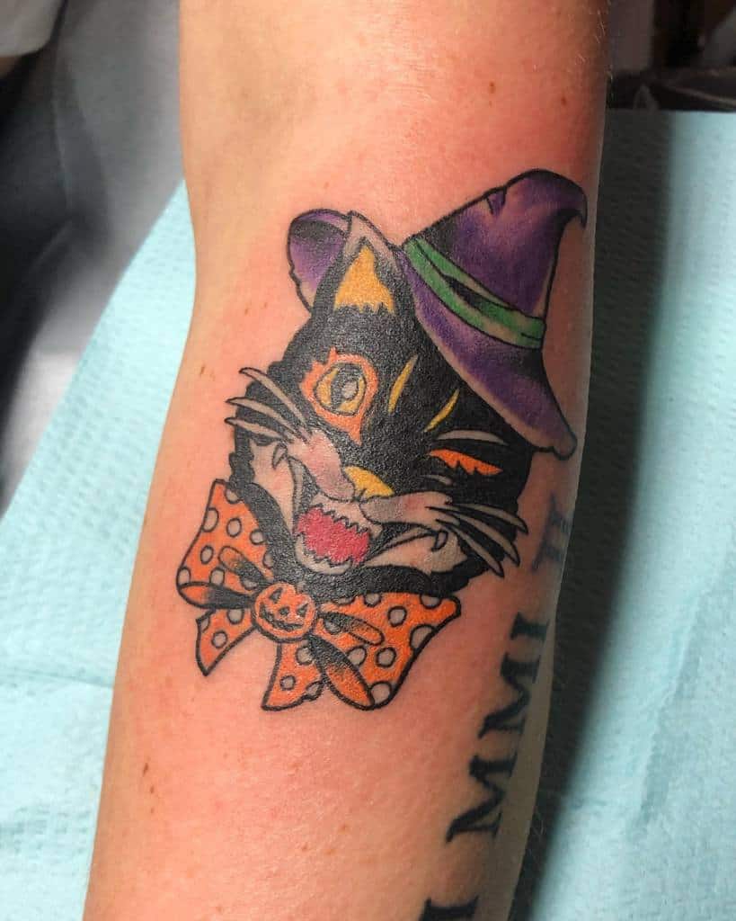 Tattoo of a charming cat donning a purple witch hat and an orange polka-dot bow tie