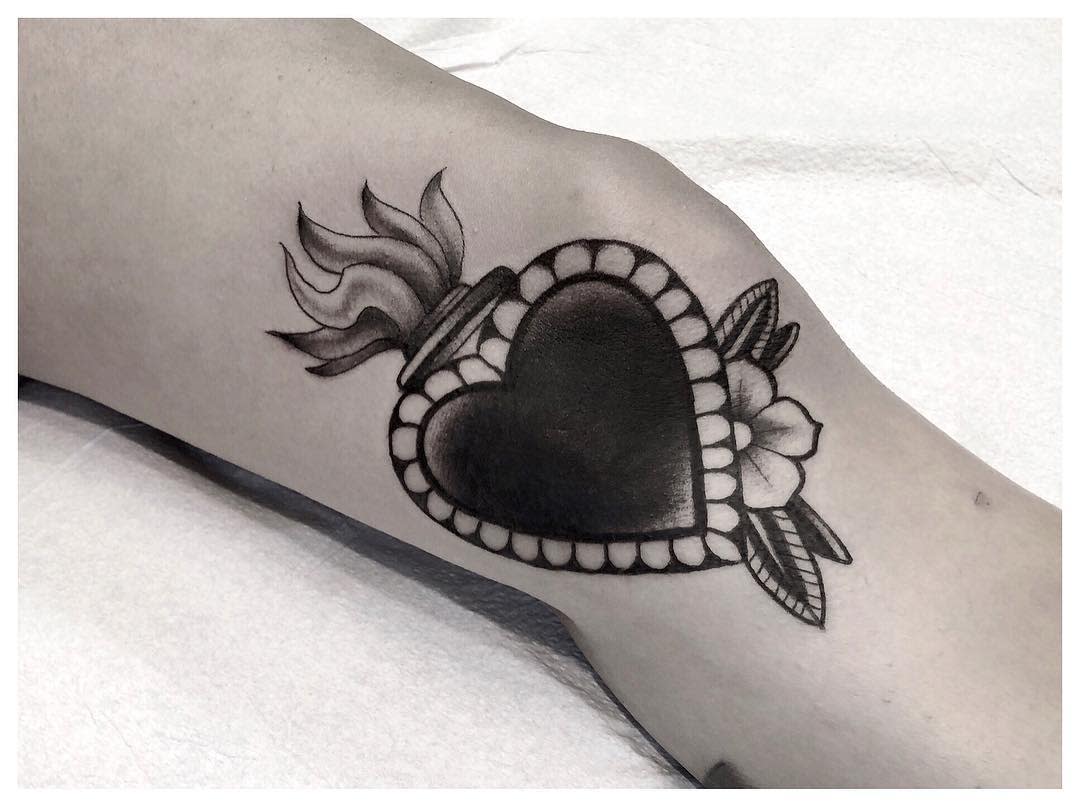 Black and grey sacred heart tattoo with bold shading, beaded border, floral details, and flame crown