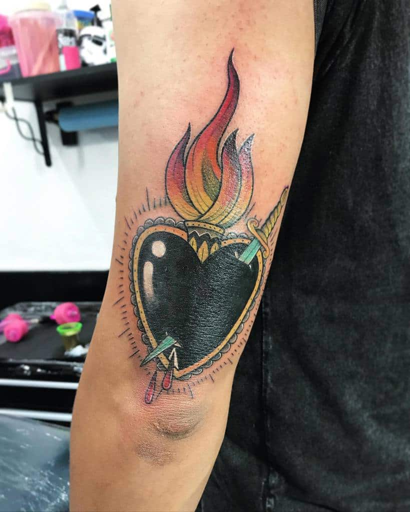 Black heart tattoo with a gold border, vibrant flames, and a dagger piercing through, surrounded by radiating lines, and two droplets of blood on the bottom
