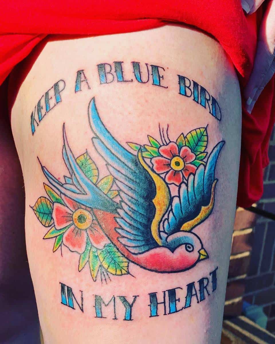 Tattoo of a blue bird surrounded by flowers with the text "Keep a blue bird in my heart" on a thigh