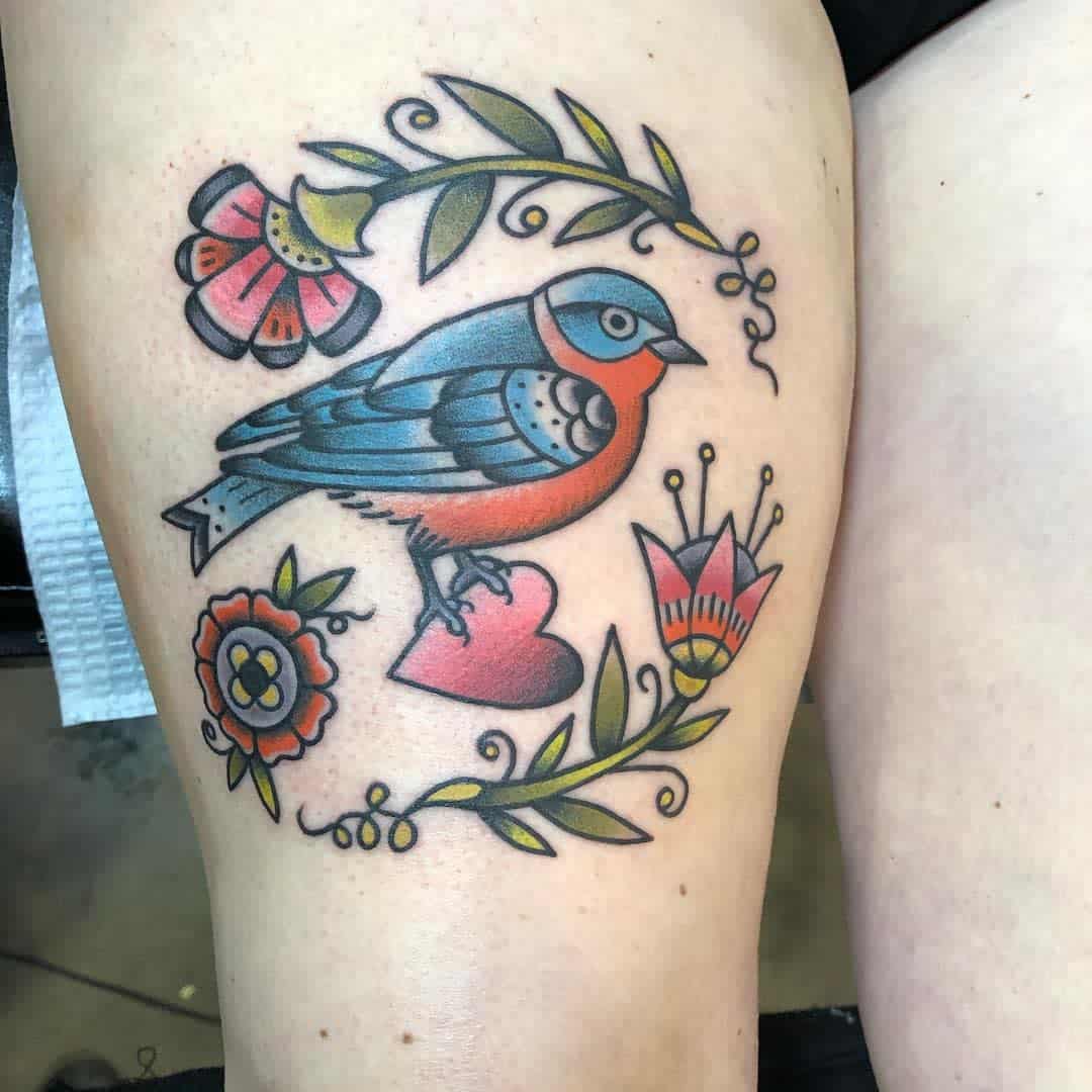 Colorful tattoo of a bird perched on a heart, surrounded by flowers and leaves on a thigh